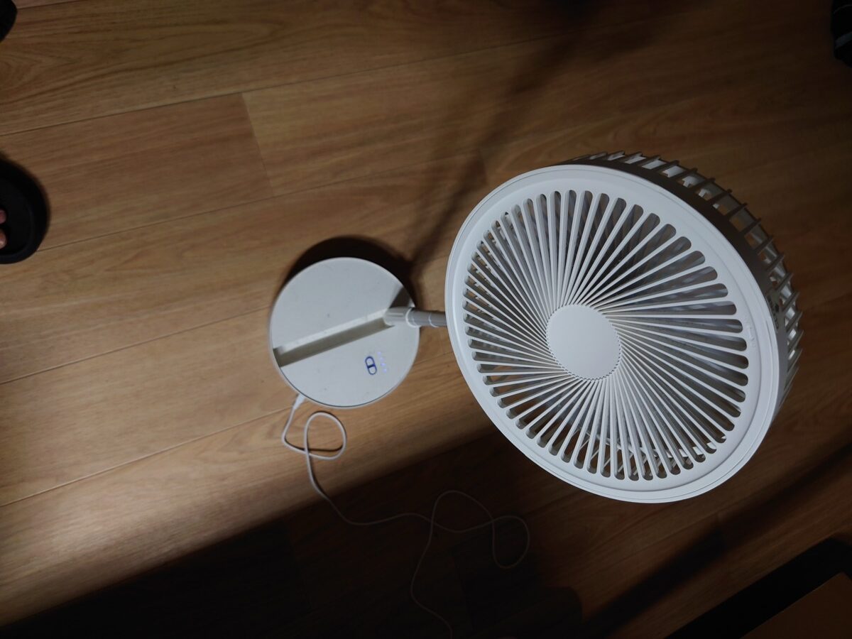 床に置いた白い折りたたみ式の扇風機を真上から見たところ。円形の台座と羽根、電源ケーブルが写っている